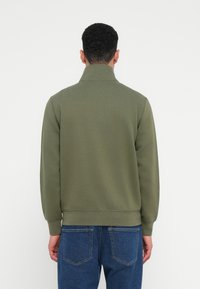 Ελαιόχρωμο φούτερ με ψηλό γιακά και ριμπά επιθέματα. Μαλακό, υφασμάτινο υλικό, απεικονίζεται από πίσω, σε συνδυασμό με μπλε τζιν.