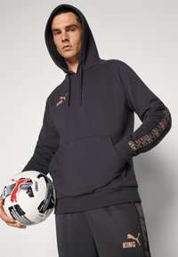 Donkergrijze Puma-hoodie met een kangoeroezak, met de tekst "KING" in koperkleur, geometrisch patroon op de mouwen, gecombineerd met bijpassende joggingbroek en een bal vastgehouden.