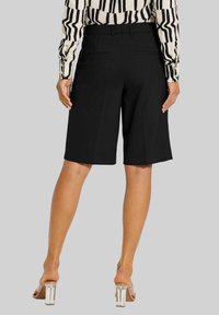 Schwarze maßgeschneiderte Shorts mit geradem Schnitt, mittlerer Oberschenkel-Länge und Gesäßtaschen. Kombiniert mit einer gemusterten Bluse mit langen Ärmeln und klaren Absätzen.