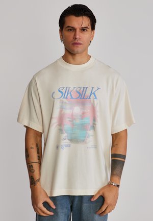 Mann trägt ein cremefarbenes, übergroßes T-Shirt mit buntem tropischem Sonnenuntergang und der Aufschrift "SIKSILK", kombiniert mit blauen Jeans, vor einfachem Hintergrund.