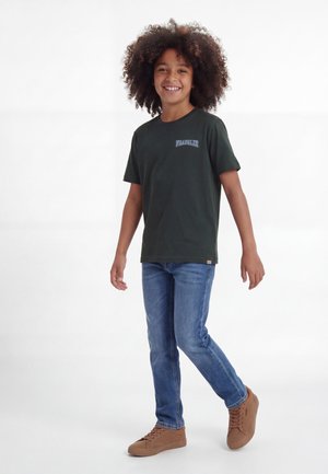 Uśmiechnięte dziecko z kręconymi włosami idące, ubrane w czarną koszulkę Wrangler, niebieskie dżinsy i brązowe trampki na białym tle.