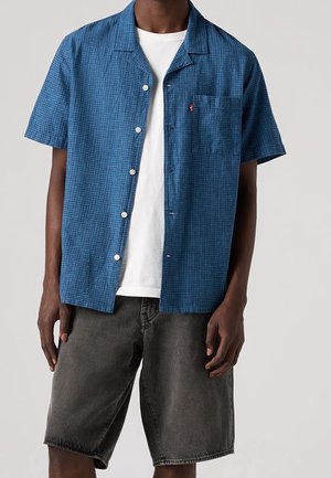 Hombre con una camisa azul texturizada de manga corta abotonada sobre una camiseta blanca y pantalones cortos de mezclilla gris oscuro sobre un fondo liso.