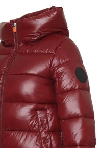 Giacca a compressione rossa con colletto alto, chiusura a zip frontale e una discreta toppa con logo sulla manica sinistra. Texture lucida e trapuntata.