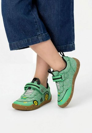 Kind trägt grün-gelbe Sneakers mit braunen Sohlen und Riemen, kombiniert mit dunkelblauen, gekürzten Hosen.