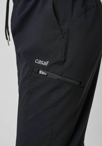 Sorte træningsbukser med en struktureret overflade, der har en lynlåslomme og "casall" logoet broderet i hvidt. Snor i taljen synlig.