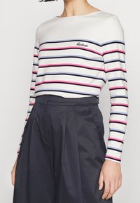 Weißes Langarmoberteil mit horizontalen schwarzen, blauen und pinken Streifen; runder Halsausschnitt; gesticktes Logo auf der linken Brust. Kombiniert mit einem dunklen Rock.