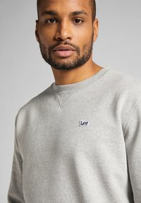 Harmaa fleece-paita, jossa on pyöreä kaula-aukko, ribattu hihansuu ja helma, ja rintakehässä pieni sininen "Lee"-logo. Sileä pinta.