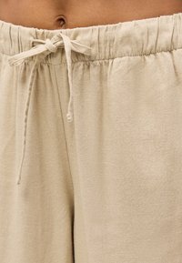 Κοντινό πλάνο σε μπεζ παντελόνι με ελαστική μέση και δεμένο κορδόνι στη μέση ενός ατόμου, με ορατό δέρμα πάνω από τη γραμμή της μέσης.