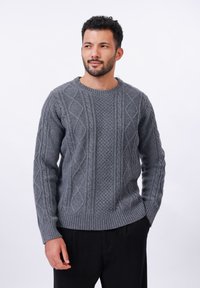 Pull en maille gris avec des motifs en diamant texturés, col rond et bords côtes aux poignets et à l'ourlet. Porté avec un pantalon noir.