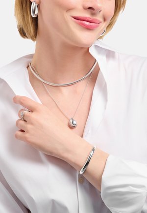 Frau mit silbernen Creolen, einer dicken Choker-Halskette, einer Tropfen-Anhänger-Halskette, einem silbernen Armreif und einem Ring mit klarem Stein am Finger.
