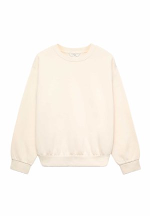 Femme aux longs cheveux bouclés porte une veste beige, un pull écru, un jean bleu et des baskets marron, debout les mains sur les hanches.