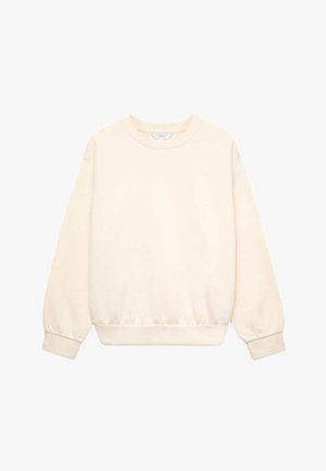Mujer con cabello largo y rizado que lleva una chaqueta beige, un suéter blanco roto, vaqueros azules y zapatillas marrones, de pie con las manos en las caderas.