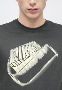 Čierne bavlnené tričko s veľkým svetlým logom "Nike" s zaoblenými okrajmi a tieňovými efektmi na jednoduchom pozadí.