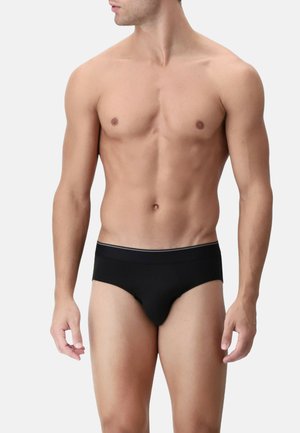 Modello maschile che indossa slip neri, in piedi con le braccia rilassate, su uno sfondo chiaro e semplice.