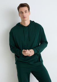 Hanorac de culoare verde închis, fabricat dintr-un material moale, dotat cu un buzunar frontal, un glugă ajustabilă cu șnur și un logo subtil pe piept. Croială slim.