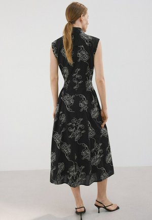 Mujer con cabello castaño recogido en una cola de caballo, vistiendo un vestido midi negro sin mangas con estampados florales blancos y sandalias negras con tacón, de espaldas.