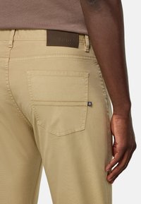 Pantalones beige con un bolsillo trasero, etiqueta de cuero estampada con "BOGGI MILANO" y un acabado de tela suave y texturizada.