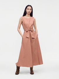 Femme portant une longue robe sans manches rose poussiéreux avec un large nœud à la taille et des bottines marron, debout les mains dans les poches.