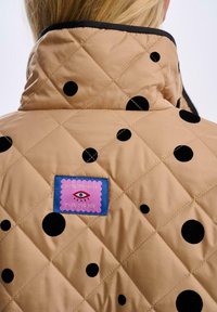 Prošívaná béžová bunda s velkými černými puntíky, vysokým límcem a barevnou nášivkou s vyšitým textem a obrázkem oka.