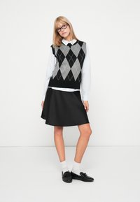 Fekete és szürke argyle kötött mellény fehér galléros ing fölött, fekete A-vonalú szoknyával és fekete, masnival díszített loaferekkel párosítva.