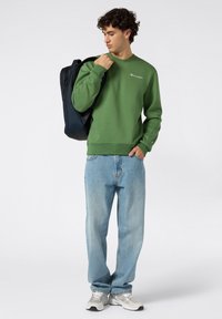 Sudadera verde con cuello redondo y logotipo, combinada con jeans de color azul claro. Sosteniendo una mochila oscura, lleva puestos unos zapatos deportivos grises.