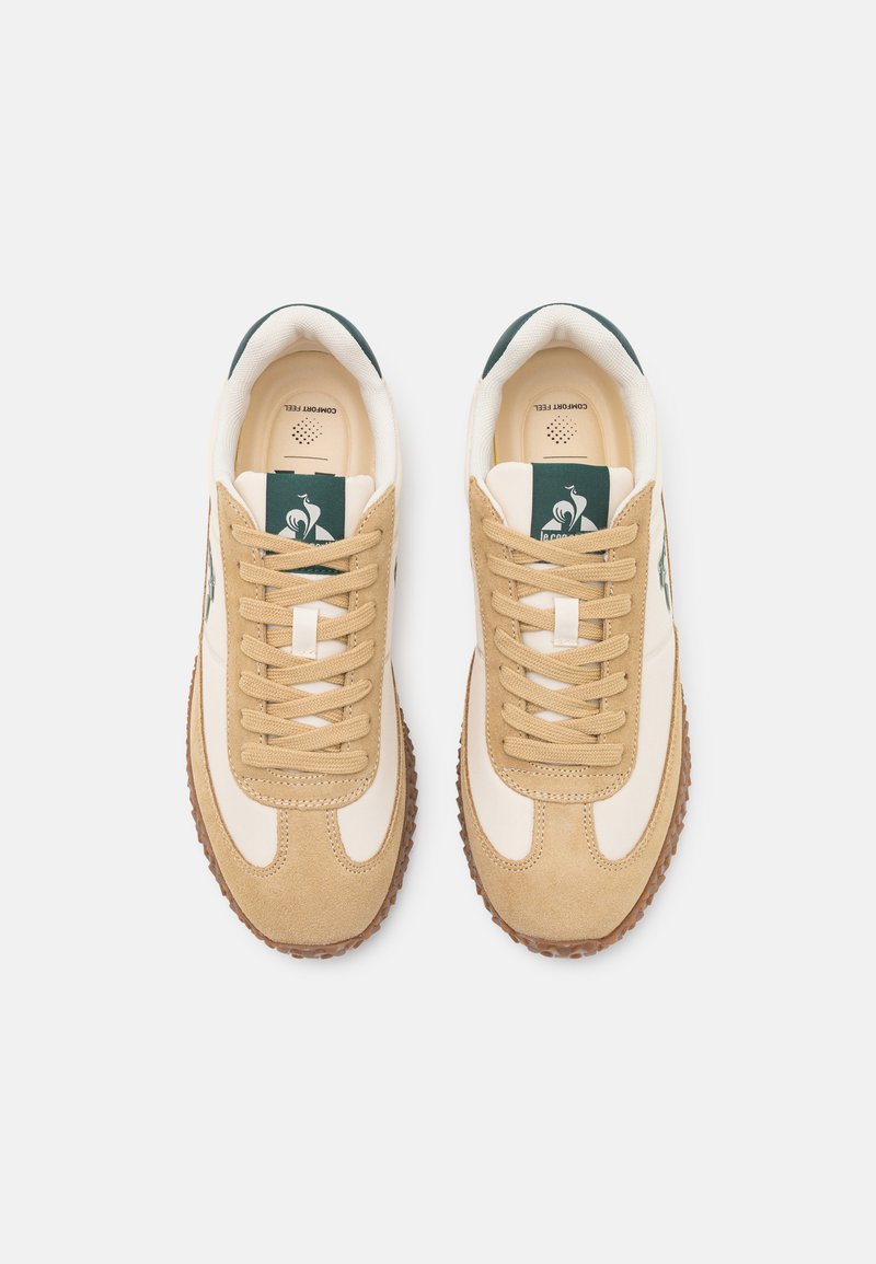 Baskets beiges et crème en daim et tissu lisse, avec un design à lacets, des semelles en caoutchouc et un logo vert sur la languette.