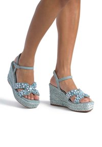Sandali con zeppa in blu chiaro, con una suola in tessuto intrecciato, dettagli in cristallo sulle cinghie e una fibbia regolabile alla caviglia.