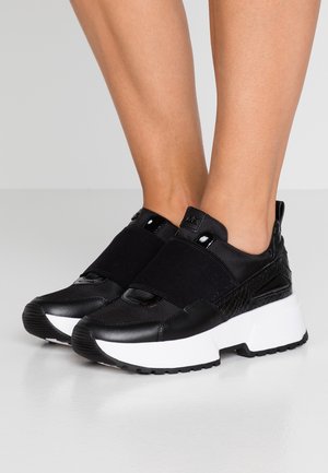 Baskets plateforme noires sans lacets avec semelles blanches épaisses portées pieds nus, avec des bandes élastiques sur le dessus et des détails texturés.