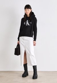 Svart hettegenser med hvit "Calvin Klein Jeans" logo, sammen med en hvit denim skjørt med frontåpning, og svarte chunky hæler støvler.