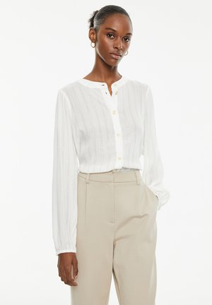 Blusa bianca a maniche lunghe con colletto abbottonato, caratterizzata da righe verticali e un leggero luccichio. Abbinata a pantaloni beige a vita alta.