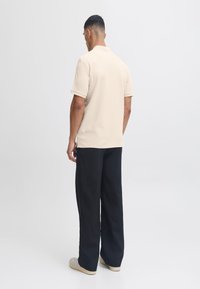 Beige polo shirt met korte mouwen, textuurstof en platte gebreide kraag, gecombineerd met loszittende zwarte broeken en schoenen met een lichte zool.