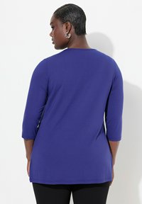 Maglia a maniche lunghe di colore viola realizzata in tessuto morbido, con scollo rotondo, maniche a tre quarti e un orlo posteriore leggermente allungato.