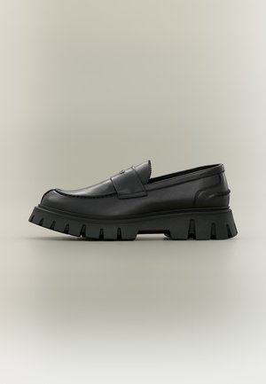 Mocassin en cuir noir avec une semelle en caoutchouc épaisse à nervures et une bride décorative sur le dessus, présenté en profil latéral sur fond neutre.