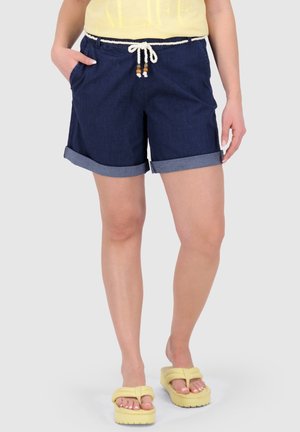 Frau trägt dunkelblaue, hochgekrempelte Jeansshorts mit weißem Kordelzug und gelben Plateausandalen, steht mit den Händen in den Taschen.