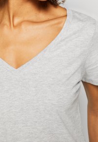 Gros plan d'une personne portant un T-shirt gris clair à col en V, montrant sa clavicule et une partie de son bras droit.