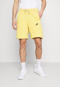 Homme portant un short jaune Nike, des chaussettes blanches et des baskets blanches, se tenant contre un fond clair uni avec une main dans sa poche.