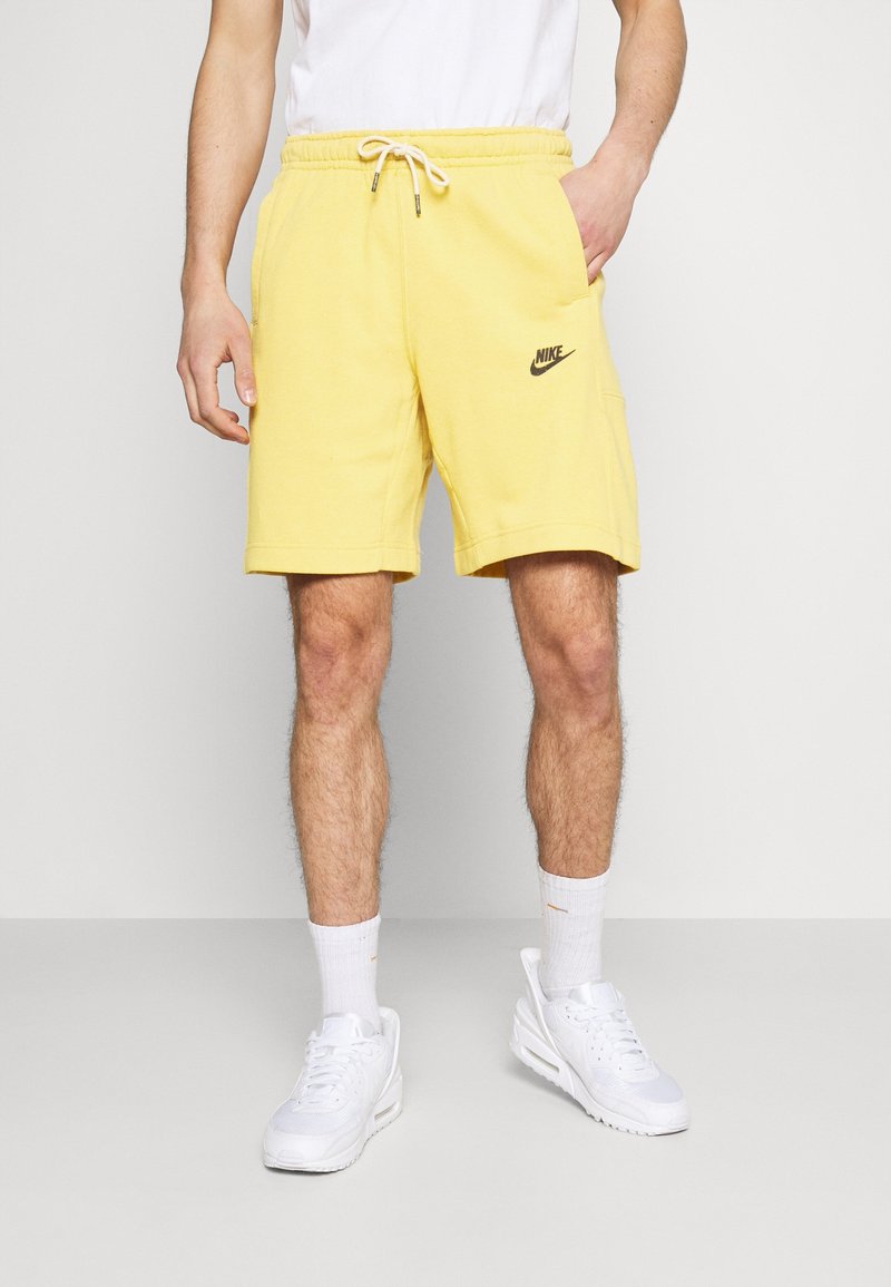 Homme portant un short jaune Nike, des chaussettes blanches et des baskets blanches, se tenant contre un fond clair uni avec une main dans sa poche.