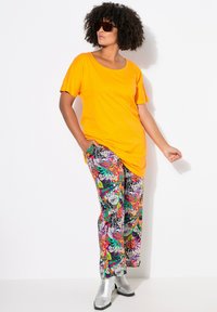 Helderoranje t-shirt, losse pasvorm, gecombineerd met kleurrijke, gemusterde broeken met bloemmotieven en tekst. Zilveren enkellaarsjes maken de outfit compleet.