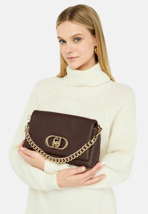 Bruine leren handtas met een gestructureerde afwerking, kettinghengsel en een goudkleurige sluiting. Het model draagt een witte coltrui.
