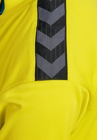 Gul sportstopp med en texturerad svart sidopanel som har ett geometriskt mönster av vita pilar och en slät tygfinish.