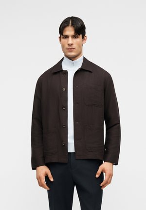 Mężczyzna ubrany w ciemnobrązową zapinaną na guziki kurtkę, pod którą ma jasnoszary top na zamek, oraz ciemne spodnie, stoi przodem na jednolitym tle.