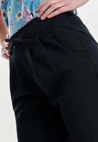 Pantaloni neri con una vestibilità comoda, vita regolabile con cordoncino, rouches davanti e tasche laterali. Il tessuto ha una finitura liscia e opaca.