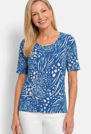 Femme blonde souriante, portant un haut à manches courtes bleu avec un imprimé animal abstrait blanc et un pantalon blanc, debout devant un fond uni.
