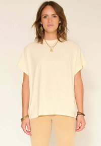 Femme aux cheveux bruns mi-longs portant un haut crème ample à manches courtes, un pantalon beige, des bijoux en or, et se tient devant un fond clair et neutre.