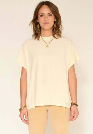 Femme aux cheveux bruns mi-longs portant un haut crème ample à manches courtes, un pantalon beige, des bijoux en or, et se tient devant un fond clair et neutre.