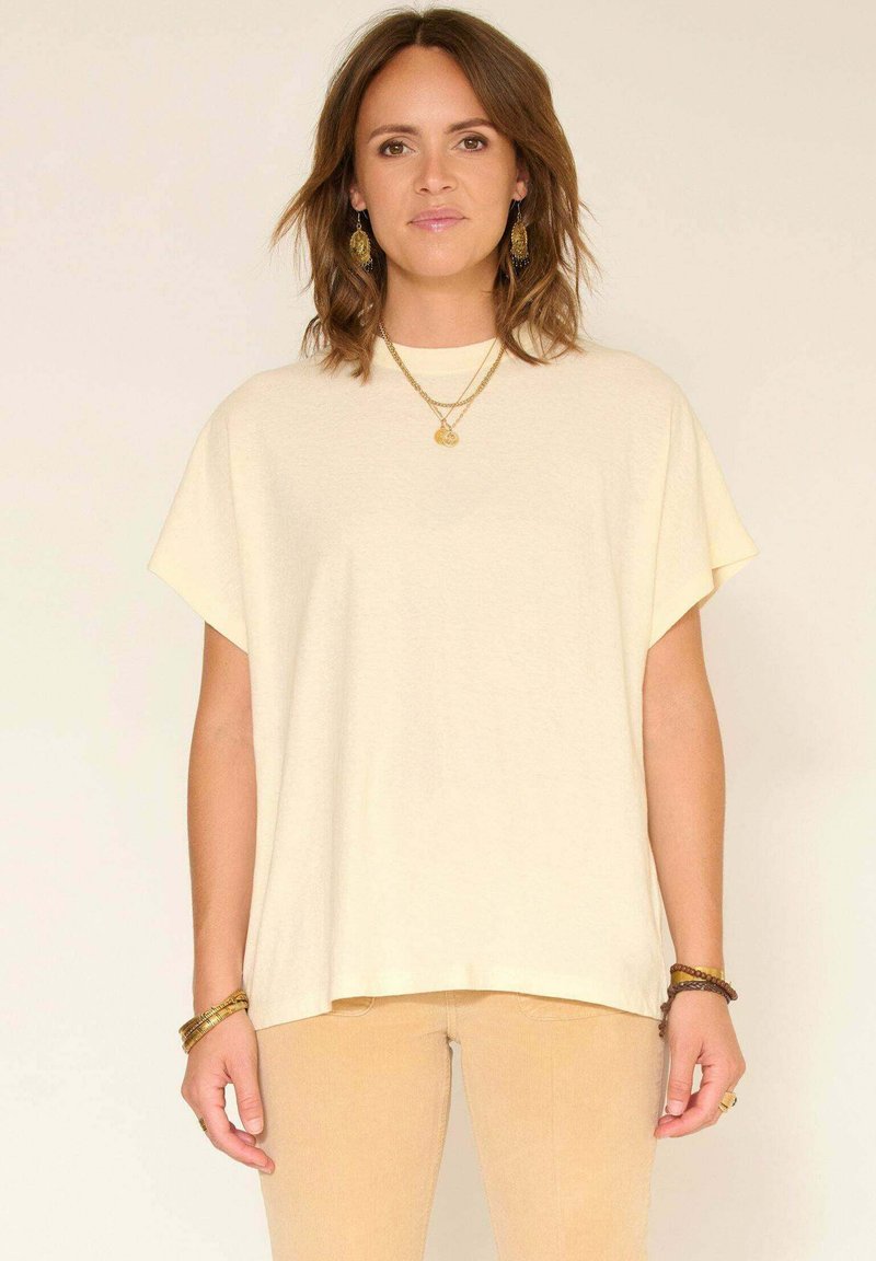 Femme aux cheveux bruns mi-longs portant un haut crème ample à manches courtes, un pantalon beige, des bijoux en or, et se tient devant un fond clair et neutre.