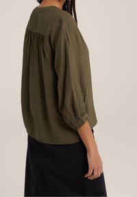 Olijfgroene, losvallende blouse met driekwart mouwen, zachte gekreukte textuur en geplooide details aan de achterkant.