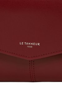 Borsa a tracolla in pelle rossa con una superficie liscia, dotata di una patta triangolare e logo in rilievo "LE TANNEUR 1898" in argento.