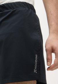 Gros plan sur la main d'une personne à côté d'un short de sport noir avec une taille élastique et le logo "Houdini" sur le côté.