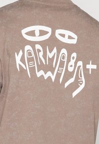 Brown t-shirt with a textured finish featuring white graphic eyes and hand designs, text reading "KARMA" in stylized lettering.